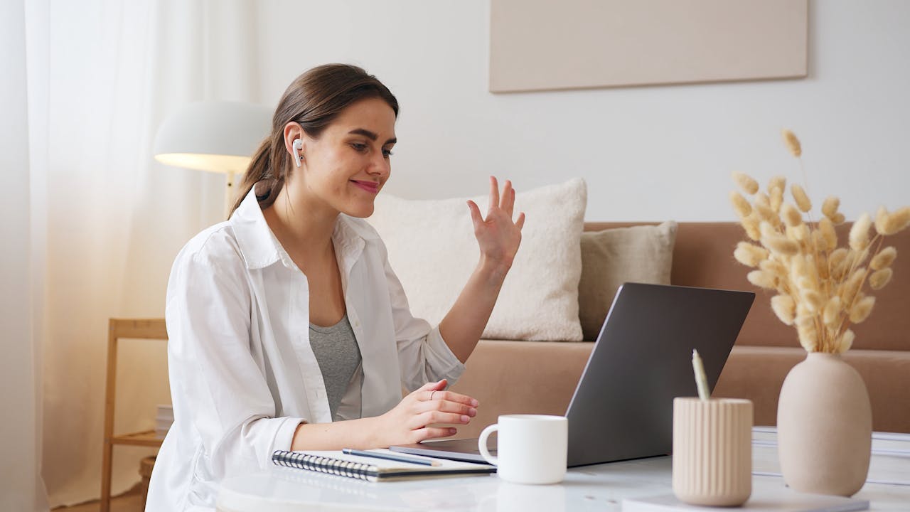 Courses Young woman attending an online class on a laptop, smiling and waving while wearing wireless earbuds – Total Educator