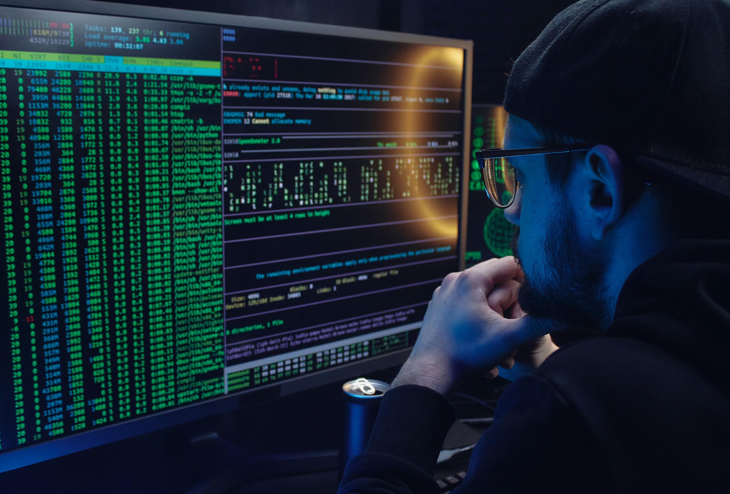 Certified Ethical Hacker Man intently working on computer programming with code displayed on dual monitors in a dimly lit room.